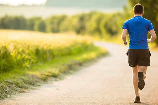 Ein sportlicher Mann läuft einen Weg entlang einer Wiese. Der Mann trägt ein blaues T-Shirt und eine kurze schwarze Hose.