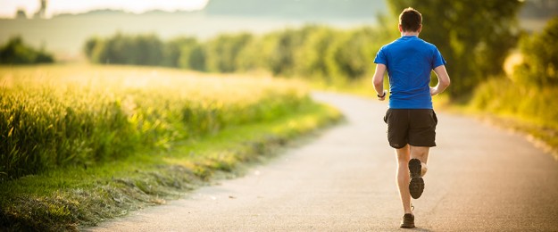 Ein sportlicher Mann läuft einen Weg entlang einer Wiese. Der Mann trägt ein blaues T-Shirt und eine kurze schwarze Hose.