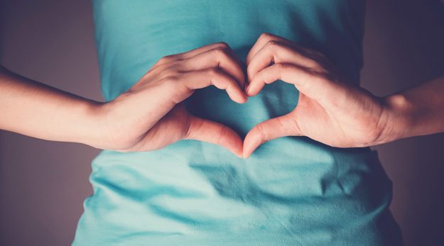 Eine Frau mit türkisfarbenen T-Shirt bildet mit ihren Händen vor ihrem Bauch ein Herz