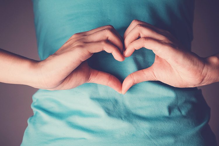 Eine Frau mit türkisfarbenen T-Shirt bildet mit ihren Händen vor ihrem Bauch ein Herz