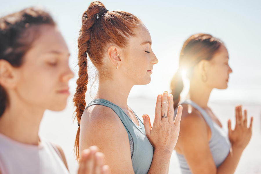 Drei Frauen machen Yoga mit geschlossenen Augen und halten die Hände im Gebet vor dem Brust.