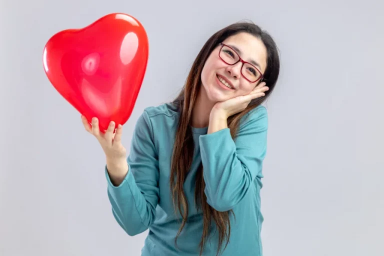 Frau im blauen Pulli hält einen roten herzförmigen Ballon und lächelt.