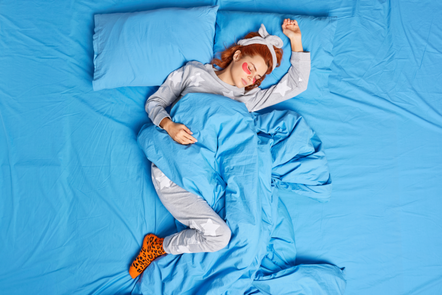Junge Frau liegt schlafend in einem großen blauen Bett. Sie trägt Schlafanzug, ein Haarband und Augenringe-Pads