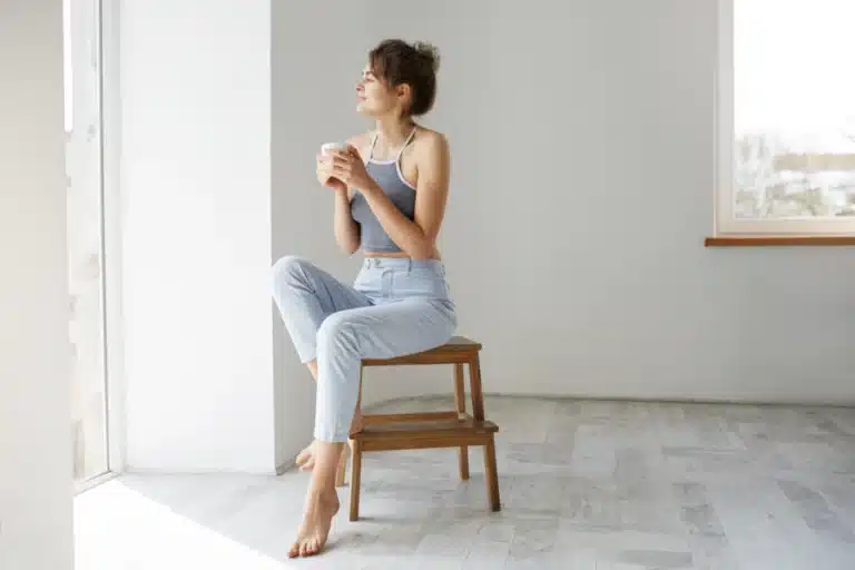 Junge Frau sitzt am Fenster auf einem Hocker und genießt am Morgen ihren Kaffee.