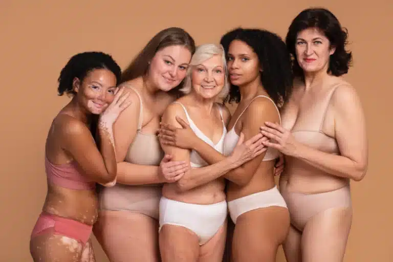 Fünf Frauen mit unterschiedlichen Körperformen, unterschiedlicher Hautfarbe und unterschiedlichem Alter liegen sich lächelnd in den Armen.