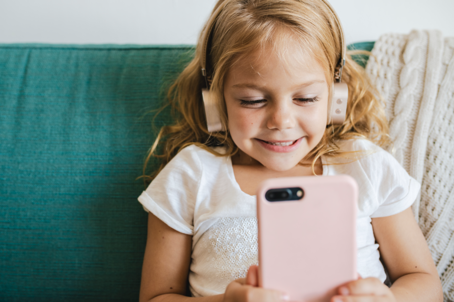 Ein kleines blondes Mädchen mit Kopfhörern schaut begeistert auf ein rosanes Smartphone.