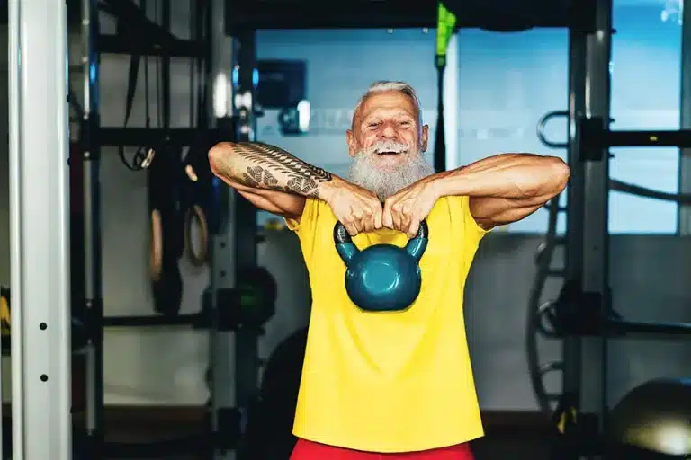 Älterer Mann im Fitnessstudio. Er trägt ein gelbes T-Shirt und hat weiße Haaren und einen längeren weißen Bart. In seinen schulterhoch gehaltenen Händen hält er ein Gewicht.