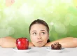 Mädchen das auf einen Tisch gestützt nachdenkt. Vor ihr stehen ein Apfel und ein Kuchen