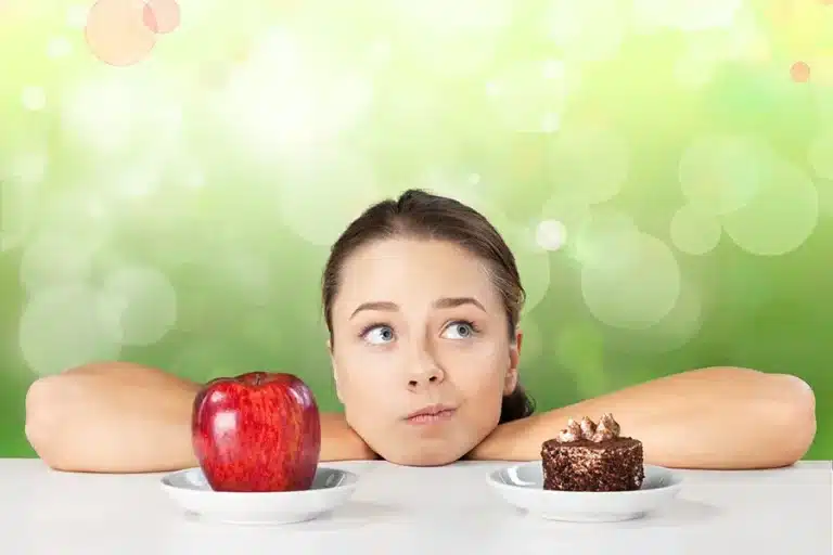 Mädchen das auf einen Tisch gestützt nachdenkt. Vor ihr stehen ein Apfel und ein Kuchen