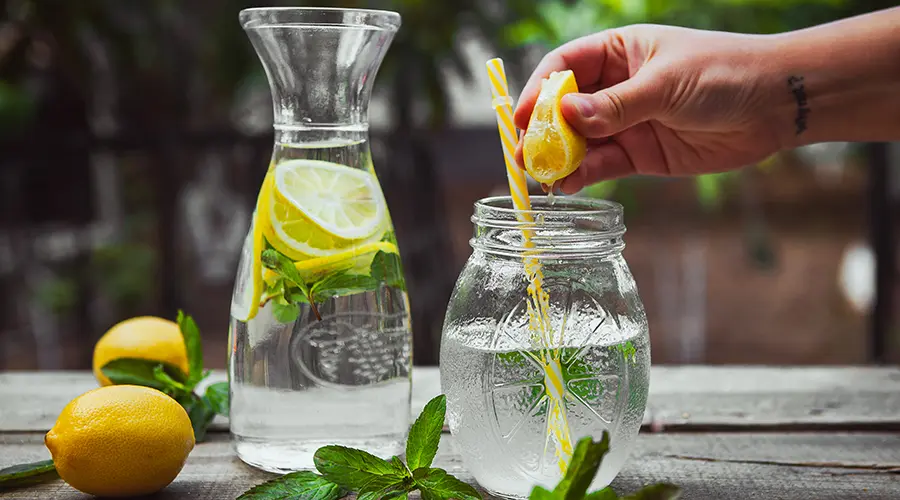 Eine Hand, die eine Zitrone in ein Glas Wasser mit Minze presst. Daneben steht eine Karaffe, die Zitronenscheiben, Minze und Wasser enthält