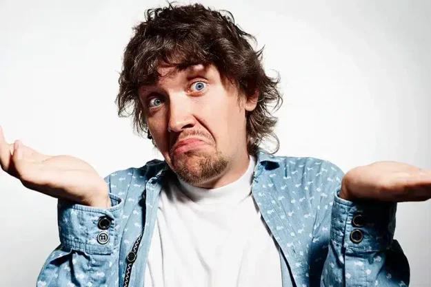 Ein Mann, im offenen blauem Hemd und weißen Shirt, der eine Schulterzuckende Geste andeutet