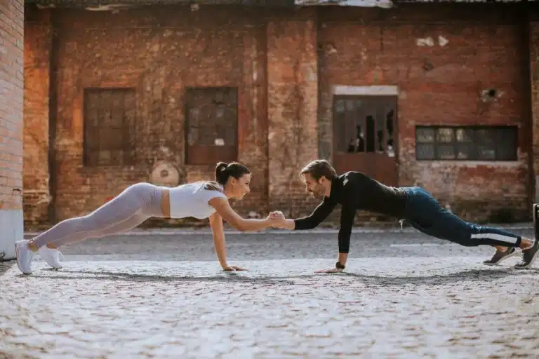 Zwei sportlich gekleidete Menschen, die Liegestütze machen und sich dabei die Hand reichen.