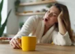 Eine gähnende Frau sitzt an einem Küchentisch. Vor ihr steht eine gelbe Kaffeetasse.